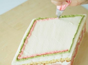 step18: 分別將食用紅色色素和抹茶粉加入表層裝飾用的鮮奶油中，打至8分發泡，製作出粉紅色和綠色的發泡鮮奶油。用格拉辛紙（glassine paper）製作擠花袋，分別將不同顏色的發泡鮮奶油裝入，擠在蛋糕表面。最後放上杏仁糖膏製作的娃娃裝飾。