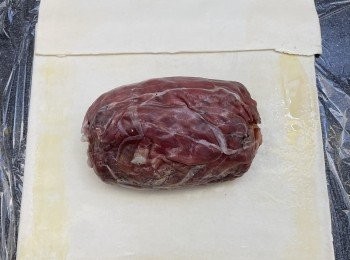 step6: 酥皮稍為軟化，量度酥皮長度，如一張不足夠包裹牛柳，需重疊酥皮，將多出嘅酥皮剪掉。工作枱面鋪上保鮮紙，酥皮放上面，酥皮面四邊掃上全蛋液。牛柳拿掉保鮮紙，牛柳放在酥皮中間，利用保鮮紙推動捲實牛柳，