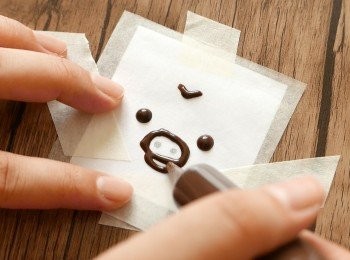 step19: 裝飾做法（步驟19-21）：
把朱古力筆浸入50°C暖水中約3分鐘至朱古力溶化。預備一張如蛋撻面大小的鼻窿雞圖樣，在圖樣上固定一張牛油紙。在牛油紙上，用黑色朱古力筆依圖樣勾畫線條。